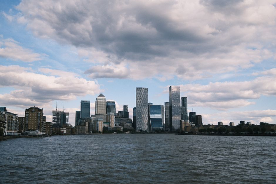 A clear view of the Canary Wharf skyline in London, featuring modern glass skyscrapers with reflective surfaces, set against a partly cloudy sky with white and grey clouds. The buildings include a mix