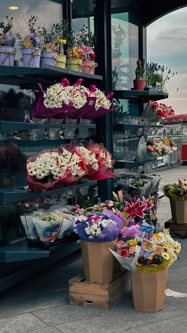 An assortment of floral arrangements and bouquets displayed on metal shelves and the pavement outside a flower shop. The top shelf holds small potted plants and wrapped flowers in pastel tones, while