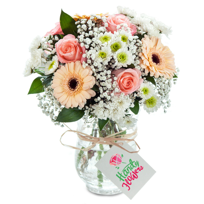 Perfectly poised on a pristine white surface, this lush floral arrangement mirrors the elegance and vitality found in London's Docklands. Central to the bouquet, velvety peach-pink roses unfurl in generous blooms, offering an immediate sense of romance and optimism along with gentle fragrance. Alongside, two impeccably shaped peach-toned gerbera daisies with striking cocoa-brown centres stand out, as bright and lively as Riverside's weekend ambience. Delicate white baby's breath clusters thread gracefully between the feature flowers, creating a feather-light volume evocative of passing clouds over Millwall Outer Dock. The composition is completed with white chrysanthemum clusters-each with an eye-catching lime-green centre-lending texture and a crisp finish. Here and there, sprigs of eucalyptus add structured greenery and a hint of the outdoors. Set within a clear, subtly textured glass vase, the bouquet's green stems can be glimpsed through water, underscoring its authenticity and vitality. Tied at the neck with a rustic raffia ribbon and adorned with a simple white tag, this arrangement stands ready for a heartfelt note. The skilled craftsmanship, gently illuminated, makes this bouquet perfect for commemorating Docklands milestones, housewarmings, or sending cheer to a friend along Marsh Wall.